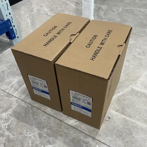 Two 3G3MX2-AB015-V2 inverter shipping cartons side by side on a tile floor, with the CAUTION HANDLE WITH CARE mark printed on the top flaps