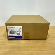 Omron C200HW-PA204S — second angle of the brown Omron-branded carton showing the same C200HW-PA204 type label, lot number 2843YK, barcode, and OMRON wordmark on a wooden tabletop