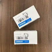 Omron CJ1W-CIF11 — two small white Omron-branded product boxes on a wooden surface, the upper box tilted and showing the CJ1W-CIF11 type label / lot number 250106K / KCC-REM-OMR-CJ1W-00061 KC mark, the lower box upright with the CJ1W-CIF11 type label and lot 250114K