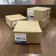 Omron CJ1W-EIP21 — brown Omron-branded shipping carton standing on a wooden surface, showing the CJ1W-EIP21 type label and the blue-and-white OMRON band along the bottom