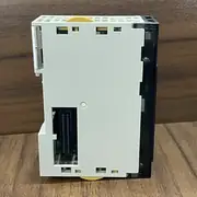 Omron CJ1W-NC413 — back / side view of the module on a wooden surface showing the white plastic housing with the yellow release lever at the top and the gold backplane connector contacts visible in the housing slot at the bottom; the silver heat-sink ventilation grid runs along the upper third of the body