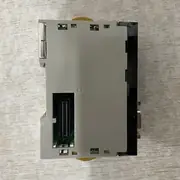 Omron CJ1W-PRM21 — underside of the PRM21 module on a wooden surface showing the gray plastic bottom housing, the gold contact fingers of the 40-pin backplane connector on the left, the D-Sub 9-pin PROFIBUS connector visible on the right edge, and the yellow release levers at top and bottom