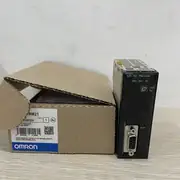 Omron CJ1W-PRM21 — open brown Omron cardboard shipping carton on a light surface showing TYPE CJ1W-PRM21 / LOT No. 240402N / QTY 1 / KCC-REM-OMR-CJ1W-00078 / barcode 4 547548 010955 / OMRON MANUFACTURING B.V. / MADE IN THE NETHERLANDS / blue OMRON band / next to the PRM21 module standing upright on the same surface