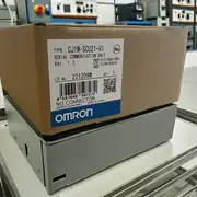 Omron CJ1W-SCU21-V1 — brown Omron-branded shipping carton resting on top of a metal-cased power supply or PSU on a white industrial surface, showing the CJ1W-SCU21-V1 type label and the blue-and-white OMRON band with warehouse electrical cabinets visible in the background