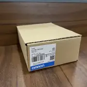 Omron CJ1W-SCU22 — small retail wrapper with white sticker label showing the CJ1W-SCU22 type code, the lot number, and the blue-and-white OMRON band, sitting on a wooden surface