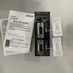 Omron CJ1W-SCU31-V1 — two black CJ1W-SCU31-V1 modules stacked vertically on a white surface next to a white Omron-branded product sheet and a pair of uninstalled 5-pin RS-422A/RS-485 terminal blocks for the COMM1 and COMM2 ports