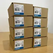 Omron CJ1W-SCU32 — eight brown Omron cardboard shipping cartons stacked in a 4-high x 2-wide grid on a wooden surface, each showing TYPE CJ1W-SCU32 / Ver.2.1 / LOT No. 230819M (one carton 180806K) / QTY 1 / KCC-REM-OMR-CJ1W-00038 KC mark / OMRON Corporation / MADE IN JAPAN / blue OMRON band