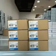 Omron CJ1W-SCU42 — stack of six brown Omron cardboard shipping cartons (3 x 2 grid) on a glass shelf showing TYPE CJ1W-SCU42 / Ver. 2.1 / lots 230303K / 221130K / 221128K / 230308K / KCC-REM-OMR-CJ1W-00039 KC mark / NO CONNECTOR note / OMRON Corporation / MADE IN JAPAN / blue OMRON band