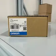 Omron CJ1W-SCU42 — single brown cardboard box at angle on a white shelf showing TYPE CJ1W-SCU42 / Ver. 2.1 / lot 230303K / KCC-REM-OMR-CJ1W-00039 KC mark / NO CONNECTOR note / OMRON Corporation / MADE IN JAPAN / blue OMRON band