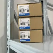 Omron CJ1W-SCU42 — three brown cardboard boxes stacked on a metal storage rack showing TYPE CJ1W-SCU42 / Ver. 2.1 / lots 230303K / 230308K / 221128K / KCC-REM-OMR-CJ1W-00039 KC mark / NO CONNECTOR note / OMRON Corporation / MADE IN JAPAN