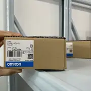 Omron CJ1W-SCU42 — single brown cardboard box held in hand showing TYPE CJ1W-SCU42 / Ver. 2.1 / lot 230308K / KCC-REM-OMR-CJ1W-00039 KC mark / NO CONNECTOR note / OMRON Corporation / MADE IN JAPAN / blue OMRON band