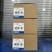 Omron CP1W-DA021 — three brown OMRON cardboard shipping cartons stacked vertically on a blue workbench with the centre of each front label reading TYPE CP1W-DA021 / PLC 模拟量输出单元 / LOT No. 05324S / QTY. 1 / KCC-REM-OMR-CP1W-0013 KC mark / barcode 4 547648 359528 / OMRON Corporation / MADE IN CHINA / blue OMRON band / small service-window sticker