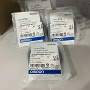 Omron E2B-M12KN05-WZ-B1 M12 inductive sensor — front view showing the M12 brass nickel-plated barrel with the PBT sensing face on the front end and the 4-conductor PVC cable exiting the rear end with brown, blue, black, and white conductors visible per IEC 60947-5-2
