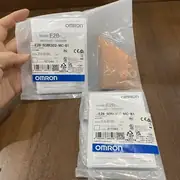 Omron E2B-S08KS02-MC-B1 inductive sensor — hand holding three sealed Omron-branded plastic bags with E2B-S08KS02-MC-B1 specification labels, one bag open showing the stainless M8 threaded sensor body inside