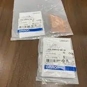 Omron E2B-S08KS02-MC-B1 inductive sensor — two sealed Omron-branded plastic bags on a wooden surface with the stainless steel M8 threaded sensor body visible inside the right bag