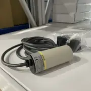 Omron E2K-C25ME1 2M — installed on a bracket facing a plastic pellet hopper inlet with the operation LED visible at the cap and the cable routed upward to the PLC input terminal