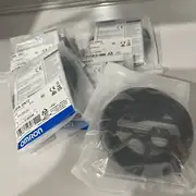 Omron E3FA-DN12 — multiple sealed anti-static clear plastic bags of E3FA-DN12 sensors stacked on a white plastic shelf, each bag showing the white E3FA-DN12 PHOTOELECTRIC SENSOR type label / 10 to 30V DC / 2m / LOT No. 01825M / QTY 1 / KCC-REM-OMR-CP1W-0013 KC mark / barcode 4 548583 312326 / OMROM Cooperation / OMRON EUROPE B.V. importer / MADE IN CHINA / blue OMRON band; the sensor body and cable visible through the bag, coil of black PVC cable in the foreground bag