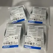 Omron E3FA-DN12 — two anti-static clear plastic bags of E3FA-DN12 sensors on a white plastic shelf with the same E3FA-DN12 / 2m / 10 to 30V DC / LOT No. 01825M labels showing the OMROM Cooperation manufacturer / OMRON EUROPE B.V. importer / KCC-REM-OMR-CP1W-0013 KC mark / blue OMRON band / barcode, with CE RoHS UKCA compliance print visible across the top of the bag