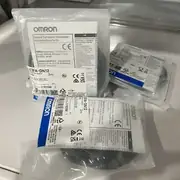 Omron E3FA-DN12 — close-up of the black M18 E3FA-DN12 sensor threaded body partly protruding from the anti-static bag in an angled view, showing the cylindrical head, the cable exit at the rear, and the white coil of cable wrapped against the body on a white plastic shelf