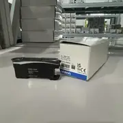 Omron E3NX-CA6 — black E3NX-CA6 fiber amplifier next to its white Omron-branded product box labeled TYPE E3NX-CA6 PHOTOELECTRIC SENSOR VOLTS 10 to 30 V DC LOT No. 22425A with the OMRON wordmark and CE / UKCA marks on a warehouse shelf