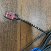 E3T-FD11 installed in a tight bracket on a feeder track showing the 12 mm thin profile of the side-view body, the 2 m cable landed on a PLC input terminal, and the orange operation LED lit when the part is present