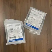 Two sealed Omron E3Z-D62 retail bags laid side by side on a wooden table, both showing the E3Z-D62 product label and Omron blue brand stripe
