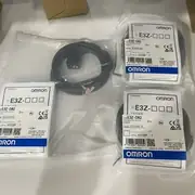 Omron E3Z-D82 diffuse-reflective photoelectric sensor — installed on a small conveyor with the visible red emitter spot on a cardboard carton 0.7 m away, M8 4-pin connector mated to an XS3F-M422-402-A PUR robotic cable returning to a CP1W-AD042 input