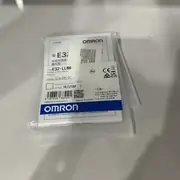 Omron E3Z-LL86 installed in a fixture with the visible red laser spot aimed at a small connector pin on an assembly jig and an M8 patch cord trailing to the panel