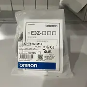Omron E3Z-T61A-M1J 0.3M — sealed anti-static bag containing the OMRON instruction booklet (showing 型 E3Z-□□□ / 光电传感器 / 使用说明书 / STANDARD), the white TYPE E3Z-T61A-M1J 0.3m / PHOTOELECTRIC SWITCH / 12 to 24V DC / LOT No. 18222M sticker, and the blue-and-white OMRON wordmark band at the bottom of the sticker, on a metal surface with other Omron carton stacks in the background