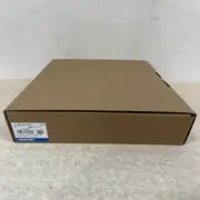 Omron NB10W-TW01B HMI — side view of sealed brown shipping box on the floor with Omron label showing 可编程控制终端 (Programmable Terminal), TYPE NB10W-TW01B, VER 1.1, KCC-REM-OMR-NB-00007 certification, LOT 05924M, QTY 1, OMRON Corporation, MADE IN CHINA, OMRON blue band