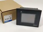 Omron NB3Q-TW01B-V1 — front view of the compact black 3.5-inch NB-series touch HMI showing the 320 x 240 color TFT display and the OMRON wordmark on the lower bezel, mounted on a wooden surface next to a brown Omron-branded product box with the NB3Q-TW01B type label