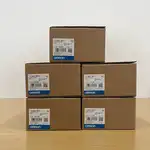 Omron NB3Q-TW01B-V1 — stack of four brown Omron-branded product boxes (2x2 grid) each with the same white NB3Q-TW01B type label, the OMRON Corporation wordmark, and the barcode label on a wooden surface