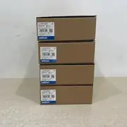 Omron NB7W-TW01B-V1 — stack of four brown Omron-branded product boxes (one on top of three) on a wooden surface, each with the white NB7W-TW01B-V1 type label, the Ver. 1.0 subtext, the KCC-REM-OMR-NB-00013 KC mark, the lot number 18125M, the barcode 4549734784597, the KC logo, and the blue-and-white OMRON band