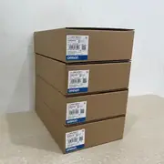 Omron NB7W-TW01B-V1 — tall stack of four brown Omron-branded product boxes on a wooden surface with a plain wall behind, each with the NB7W-TW01B-V1 type label, the Ver. 1.0 subtext, the KCC-REM-OMR-NB-00013 KC mark, the lot number 18125M, and the blue-and-white OMRON band
