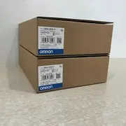 Omron NB7W-TW01B-V1 — two brown Omron-branded product boxes stacked on a wooden surface, the upper box with the NB7W-TW01B-V1 type label / VER. 1.0 subtext / lot No. 18125M / KC mark / blue-and-white OMRON band, the lower box matching