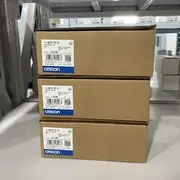 Omron NB7W-TW11B — three brown cardboard boxes stacked on a metal storage rack (lots 02320M / 11420M / 07320M), each showing TYPE NB7W-TW11B / VER. 1.1 / KCC-REM-OMR-NB-00002 KC mark / OMRON Corporation / MADE IN CHINA / blue OMRON band