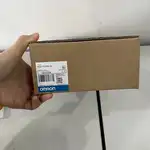 Omron NS5-TQ10B-V2 — second angle of the brown Omron-branded product box with the white NS5-TQ10B-V2 type label, the OMRON Corporation wordmark, and the barcode label, a hand holding the top corner of the box on a wooden surface