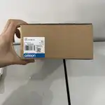 Omron NS5-TQ10B-V2 — third angle of the brown Omron-branded product box showing the top surface with the NS5-TQ10B-V2 type label and the OMRON Corporation wordmark, a hand resting on top of the box on a wooden surface
