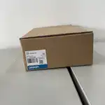 Omron NS5-TQ10B-V2 — angled view of the brown Omron-branded product box showing the side surface with the blue-and-white OMRON band, the white NS5-TQ10B-V2 type label, and the barcode label sitting on a warehouse metal shelf